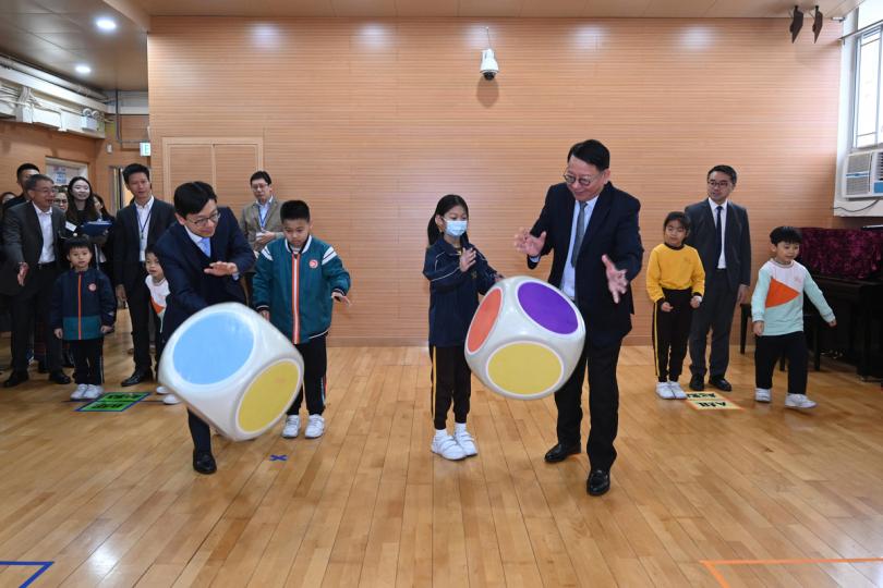 分享樂趣:  陳國基（前排右一）、孫玉菡（前排左一）了解在校課後託管服務計劃的運作情況，又與學童玩遊戲，鼓勵他們紓緩學習壓力。