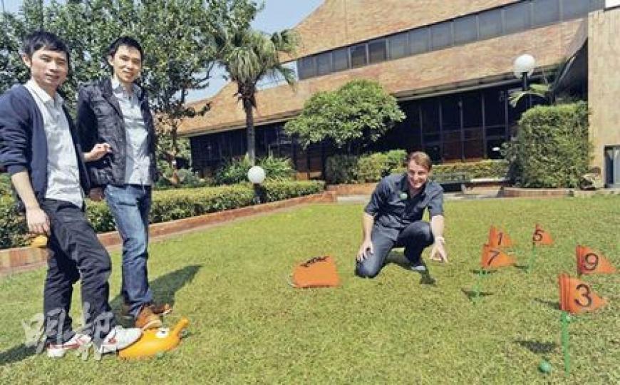 理大設計學院畢業生徐丹青（左起）、胡家禮及Chris Scolari示範奪獎玩具，玩家用手或腳按壓「發射器」（圖中橙色玩具）發球，若球降落於插有旗幟的沙洞便可得分。（尹錦恩攝）