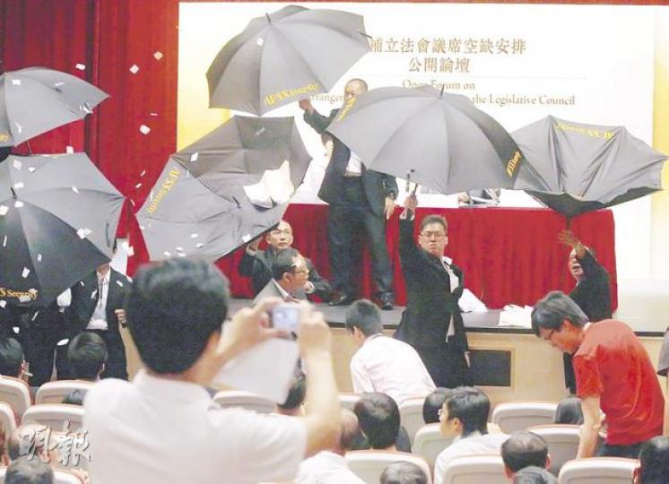 有出席論壇市民情緒激動，保安員早有準備，當有人向台上擲紙，便齊齊舉傘築起保護牆。（李紹昌攝）遞補機制首場公開論壇上，場面混亂，有示威者更將諮詢文件撕毁並擲向政制及內地事務局長林瑞麟（台上右方），幸得保安員舉傘保護，但保安員就被誤中副車。（李紹昌攝）
