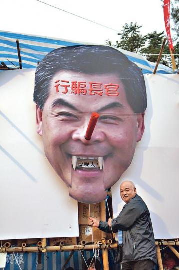 社民連攤位整咗個巨型梁振英布景，唔止個鼻會伸長，就連個嘴都郁得，一拉就會伸出「你呃人」三隻字，「阿牛」曾健成仲親身示範（圖），睇嚟社民連嘅勞作技術真係愈來愈出神入化。（曾安迪攝）