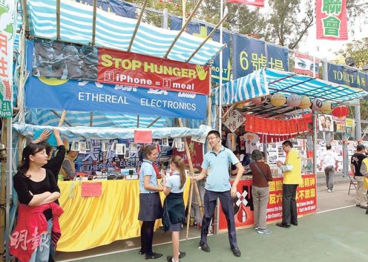維園2號攤檔（左方）現由國際學校售賣手機套作慈善籌款，前天掛在檔口的「天際數碼」橫額，被另一寫上英文標語（停止飢餓  1個iPhone=1餐飯）及饑民照片的橫額蓋着。攤檔昨日有數名穿便服成年男女（包括左一黑衣女子）看檔，3名身穿國際學校校服的學生（藍衣者）則在檔前宣傳拉客。右為支聯會的1號攤位。（李澤彤攝）支聯會今年在維園只有一攤檔，繼續主打常委張文光（左三）等人寫揮春，主席李卓人（左二）期望籌得230萬元，以興建六四永久紀念館。（李澤彤攝）