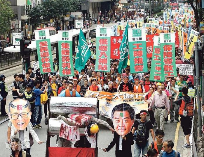 職工盟昨發起五一大遊行，遊行主題為「公平、尊嚴、家庭友善」，由正在罷工的碼頭工人打頭陣。他們在象徵官商壓榨工人的「人肉榨汁機」「帶領」下，由維園遊行至政府總部及長江中心，再前往和記大廈抗議。（李紹昌攝）