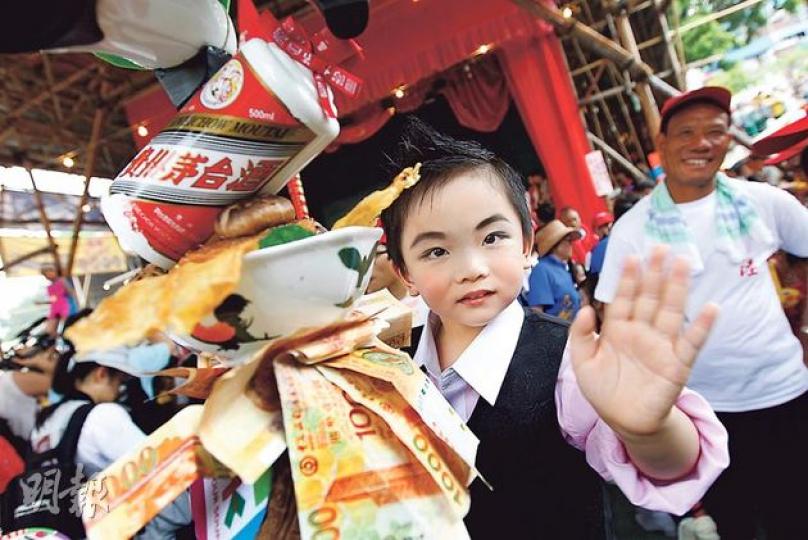 昨日太平清醮飄色巡遊打頭陣的小「湯顯明」，伴着茅台和乾鮑等裝飾，受到大批市民追捧更拍照留念。真正的湯顯明則於今日現身立法會，接受議員質詢。（李紹昌攝）參與飄色巡遊的小「湯顯明」，原定向中聯辦副主任林武（中）等嘉賓「送曲奇」，諷刺湯顯明以公帑向內地官員送禮，但這環節被大會臨時腰斬。（李紹昌攝）