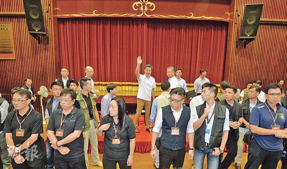 特首梁振英（後排中間揮手者）昨到觀塘與市民見面，在論壇將近完結時，人民力量在會場後方叫喊，令在場保安及警方、包括保護要員組十分緊張，隨即在台前築起三重防線，以防混亂；場內保安部署，遠較上周天水圍論壇嚴密。（盧翊銘攝）3名帶濃厚口音長者，手持寫有「818網民自發『撐CY 撐警察 護法治』宣示集會」及挺政府口號的「貓紙」，耐心聽戴黑帽女子（右）講解。長者稱自發出席，在女子手勢示意下，否認有收取車馬費和有專車接送。（張煒明攝）