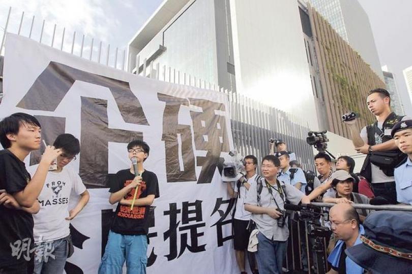 學民思潮黃之鋒（左三）昨早把「企硬公民提名」橫額掛在特首辦閘外，要求特首梁振英回應民意 ，警員即指此舉可能觸犯《公眾衛生及市政條例》，黃反駁：「我唔明呢張10呎乘10呎、白底黑字嘅banner，有咩咁唔衛生？」（李澤彤攝）特首梁振英（車廂內，左）座駕昨早7時45分抵達特首辦，但在警方沿途高舉「黃旗」護送下，梁座駕直接駛入特首辦，沒停下來與在場市民對話。（林俊源攝）