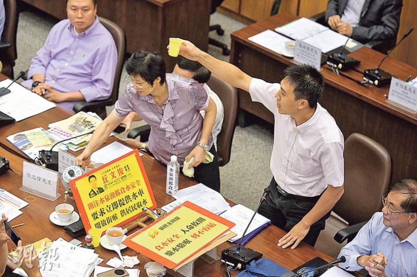 新民主同盟沙田區議員丘文俊（站着舉杯者）在區議會上向官員送上樽裝水，聲稱取自該黨驗出水泉澳邨的含鉛超標單位，他更指自己「先飲為敬」，即場喝掉，但最終只有到訪區議會的署理天文台長劉心怡有喝。（鄧宗弘攝）
