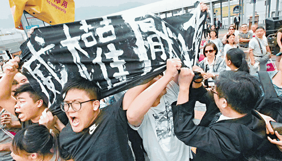 ■眾志成員在升旗期間舉起「威權壓境」橫額，遭保安扯走。林健恆提供圖片■中年漢蹲下向正被保安抬走的示威者吐口水。